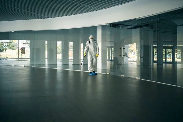 A man wearing disinfection suit spraying with sanitizer the glass doors\' handles in an empty shopping mall to prevent covid-19 spread. Health awareness, clean, defence concept