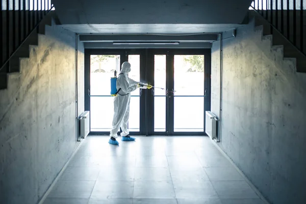 A man wearing protective disinfection suit and a spray stands in front of a glass doors under the staircases. Worker cleaning up the business center. Healthcare, covid-19 concept