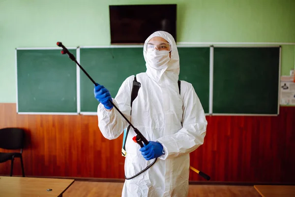 Close up of a man wearing a disinfection protective suit cleans the classroom before lessons at school. Sanitary worker sprays the auditorium. Health care of students and pupils concept