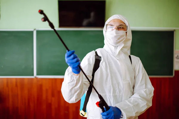 Close up of a man wearing a disinfection protective suit cleans the classroom before lessons at school. Sanitary worker sprays the auditorium. Health care of students and pupils concept