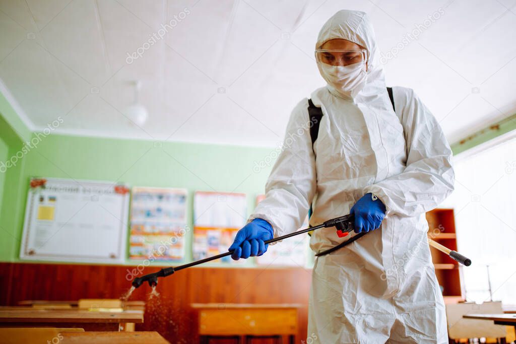Close up of a man wearing a disinfection protective suit cleans the classroom before lessons at school. Sanitary worker sprays the auditorium. Health care of students and pupils concept