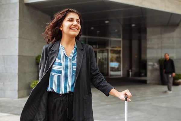 Young business woman walks with a suitcase near the hotel wearing official suit. Woman on a business trip checks out of a hotel and is waiting for a taxi. Work and travel concept