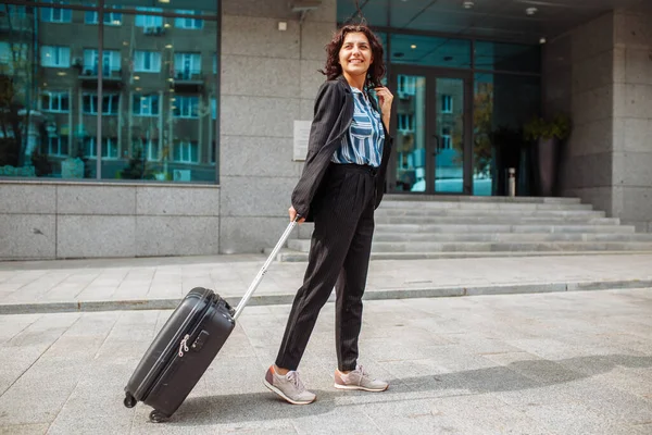 Young business woman walks with a suitcase near the hotel wearing official suit. Woman on a business trip checks out of a hotel and is waiting for a taxi. Work and travel concept