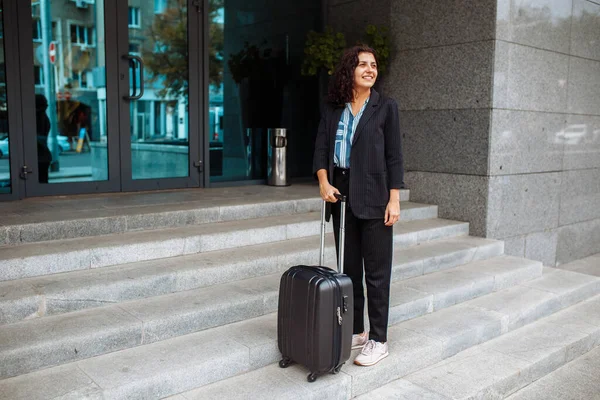 Young business woman stands with a suitcase waiting for a taxi near the hotel. Professional on a working business trip. Woman in an official suit. Work and travel concept