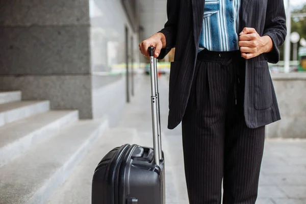 Young business woman stands with a suitcase waiting for a taxi near the hotel. Professional on a working business trip. Woman in an official suit. Work and travel concept