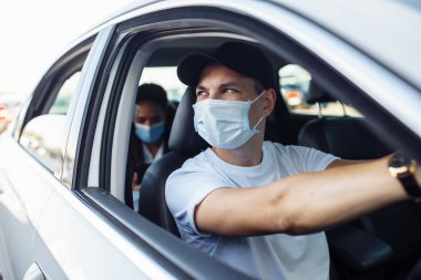 Genç bir taksi şoförü tıbbi maske takmış bir yolcuyla şehre doğru sürerken bir arabanın camından dışarı bakar. Salgın sırasında iş seyahatleri, yeni normal ve koronavirüs seyahat güvenliği kavramı