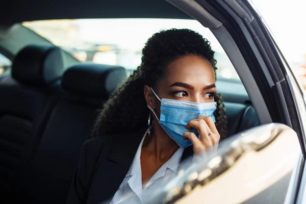 Young businesswoman being a taxi passenger and having a ride wearing and adjusting medical mask for health protection. Business trips during pandemic, new normal and coronavirus travel safety concept