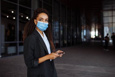 Sağlık maskesi takan genç bir iş kadını ofis merkezinin yanında duruyor. Resmi olarak elinde telefonla dışarıda bekleyen bir kız. Covid-19 salgınında önde gelen şirketler
