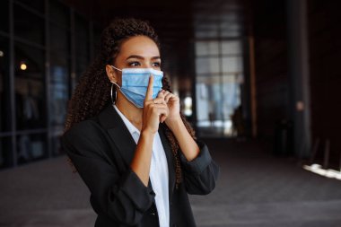 Tıbbi maske takan genç bir iş kadını cep telefonuyla konuşurken parmağının ağzının yanında sessizlik işareti gösteriyor. Covid-19 salgını sırasında bilgi eksikliği