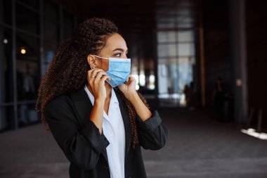 Resmi olarak takım elbiseli genç bir kadın cep telefonuyla iş konuşurken hastane maskesini ayarlıyor. Covid-19 karantina sürecinde önde gelen bir iş
