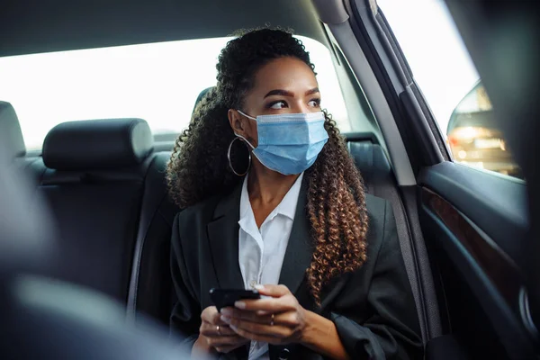 Young business woman in a mask checking her mobile cell phone on a backseat of a taxi during covid-19 quarantine. Business trips during pandemic, new normal and coronavirus travel safety concept