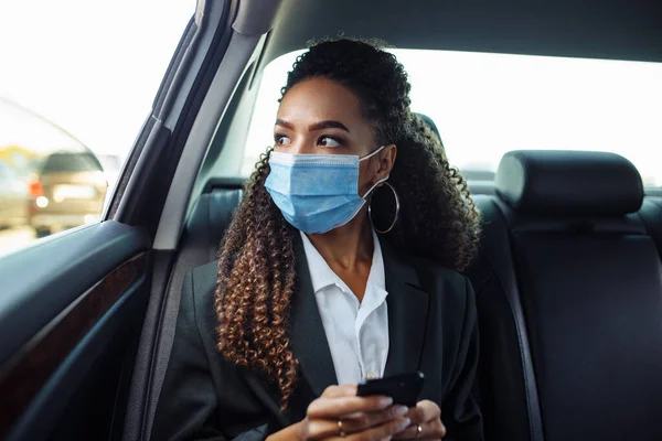 Young business woman in a mask checking her mobile cell phone on a backseat of a taxi during covid-19 quarantine. Business trips during pandemic, new normal and coronavirus travel safety concept