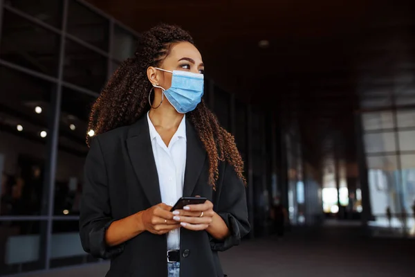 Young businesswoman wearing a medical mask stands near the office center. Officially looking girl with a phone in her hands waiting outside. Leading business during Covid-19 pandemic concept