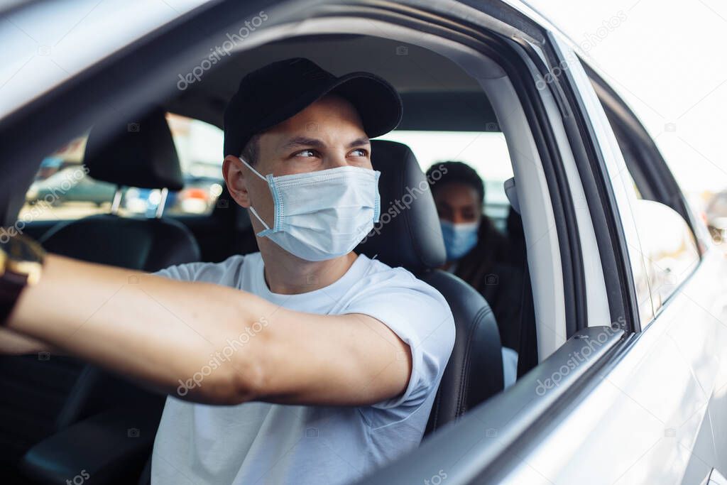 Young taxi driver looks out of a car's window while driving through the city with a passanger wearing a medical mask. Business trips during pandemic, new normal and coronavirus travel safety concept