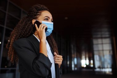 Ofis binasının dışında steril maske takarak iş konuşan genç ve çekici bir kadının portresi. Covid-19 salgınında önde gelen şirketler