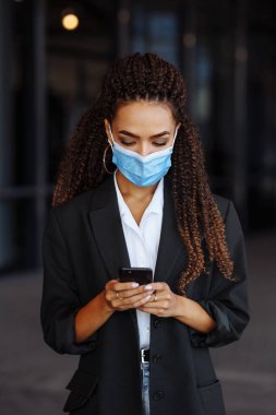 Tıbbi maske takarak cep telefonuyla mesajlaşan genç bir iş kadınının yakın çekimi. Ofisin dışında akıllı telefondan sohbet eden güzel bir kız. Covid-19 iş konsepti