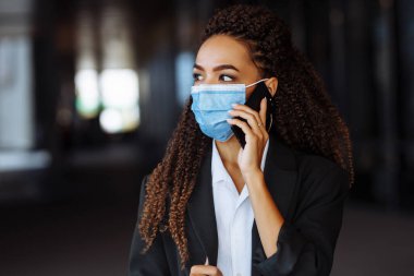 Ofis binasının dışında steril maske takarak iş konuşan genç ve çekici bir kadının portresi. Covid-19 salgınında önde gelen şirketler