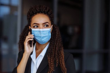 Ofis binasının dışında steril maske takarak iş konuşan genç ve çekici bir kadının portresi. Covid-19 salgınında önde gelen şirketler