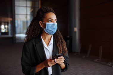Sağlık maskesi takan genç bir iş kadını ofis merkezinin yanında duruyor. Resmi olarak elinde telefonla dışarıda bekleyen bir kız. Covid-19 salgınında önde gelen şirketler