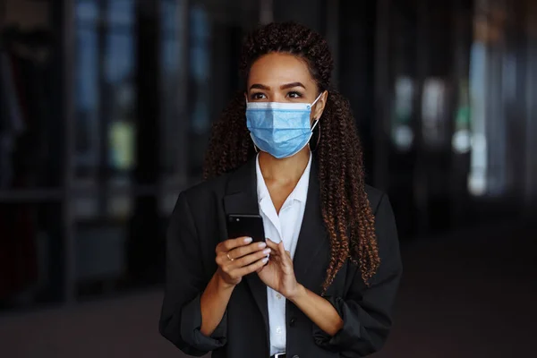 Young businesswoman wearing a medical mask stands near the office center. Officially looking girl with a phone in her hands waiting outside. Leading business during Covid-19 pandemic concept