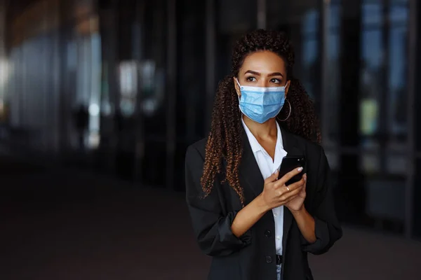 Young businesswoman wearing a medical mask stands near the office center. Officially looking girl with a phone in her hands waiting outside. Leading business during Covid-19 pandemic concept