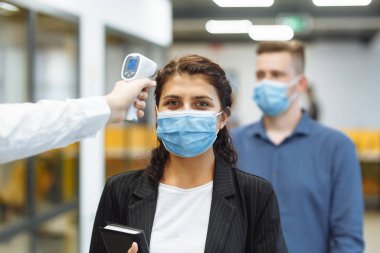 Ofis çalışanları koronavirüs salgını sırasında uzak temas olmayan bir termometreyle kontrol edilir. Ofis koridorunda sıcaklık kontrolünü geçen genç bir iş kadını covid-19 yayılmasını önlemek için.
