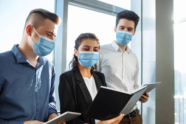 A group of collegues discuss business matters at the corridor of the office wearing medical masks to protect from coronavirus desease during global pandemic. Health safety at work concept