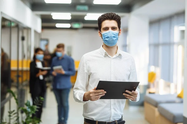 Portrait of a businessman at the office in a mask. A group of collegues discuss business matters at the corridor wearing medical masks to protect from coronavirus desease during global pandemic