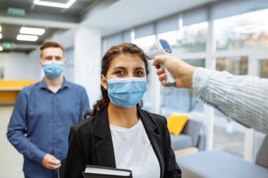 Ofis çalışanları koronavirüs salgını sırasında uzak temas olmayan bir termometreyle kontrol edilir. Ofis koridorunda sıcaklık kontrolünü geçen genç bir iş kadını covid-19 yayılmasını önlemek için.