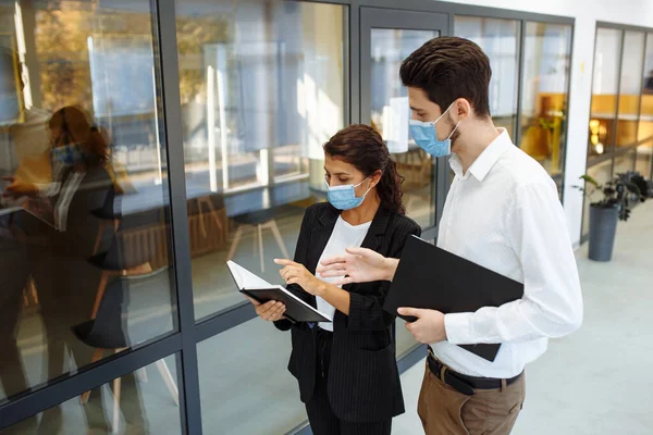 Businesspeople discuss workring issues during pandemic of coronavirus. Man and woman wearing protective medical masks talking business in the office corridor. Health safety at work concept