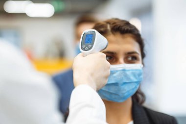Ofis çalışanları koronavirüs salgını sırasında uzak temas olmayan bir termometreyle kontrol edilir. Ofis koridorunda sıcaklık kontrolünü geçen genç bir iş kadını covid-19 yayılmasını önlemek için.