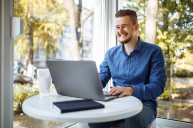 Mavi gömlekli neşeli iş adamı laptopuyla bir kafede çalışıyor. Genç adam bir fincan kahveyle mesafeli iş yapıyor. Uzak görev konsepti