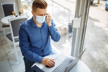 Tıbbi steril maske takan genç işadamı, bir kafeden cep telefonuyla konuşuyor, Coronavirus salgını karantinası sırasında uzaktan çalışıyor. Sağlık güvenliği kavramı