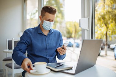 Tıbbi steril maske takan genç işadamı, bir kafeden cep telefonuyla konuşuyor, Coronavirus salgını karantinası sırasında uzaktan çalışıyor. Sağlık güvenliği kavramı