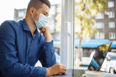 Tıbbi steril maske takan genç işadamı bir kafede dizüstü bilgisayarla oturup uzaktan çalışıyor. Uzak ve önde gelen bir işte çalışırken sağlık güvenliği. Koronavirüs konseptini yaymayı bırak