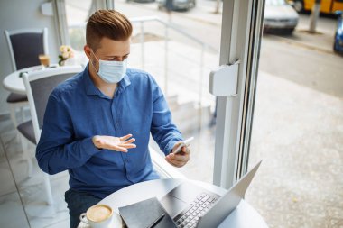 Tıbbi steril maske takan genç işadamı, bir kafeden cep telefonuyla konuşuyor, Coronavirus salgını karantinası sırasında uzaktan çalışıyor. Sağlık güvenliği kavramı