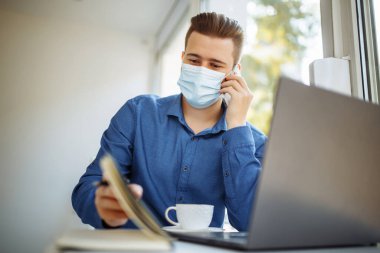 Tıbbi steril maske takan genç işadamı, bir kafeden cep telefonuyla konuşuyor, Coronavirus salgını karantinası sırasında uzaktan çalışıyor. Sağlık güvenliği kavramı