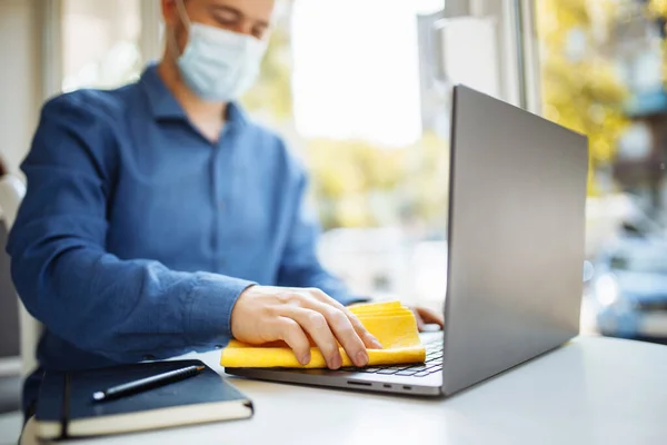 Young businessman cleans the laptop in a cafe with a yellow rag and a sanitizer to prevent coronavirus spread during worldwide pandemic. Healthcare and disinfection concept