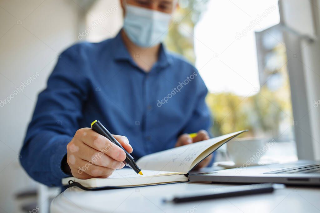 Young man taking noted and working from a cafe during coronavirus pandemic quarantine and staying safe. Leading business from a distant place. Health care concept