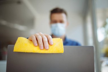 Genç işadamı, dünya çapında yayılan koronavirüsü önlemek için sarı bir bez ve dezenfektanla bir kafede dizüstü bilgisayarı temizliyor. Sağlık ve dezenfeksiyon konsepti