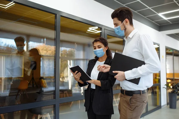 Businesspeople discuss workring issues during pandemic of coronavirus. Man and woman wearing protective medical masks talking business in the office corridor. Health safety at work concept