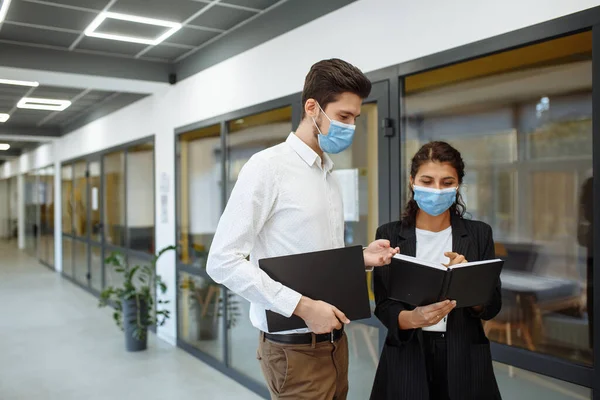 Businesspeople discuss workring issues during pandemic of coronavirus. Man and woman wearing protective medical masks talking business in the office corridor. Health safety at work concept