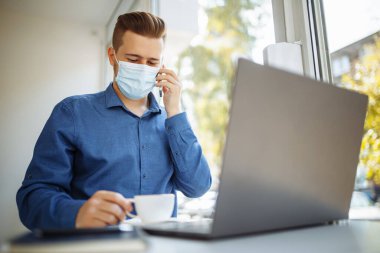 Tıbbi steril maske takan genç işadamı, bir kafeden cep telefonuyla konuşuyor, Coronavirus salgını karantinası sırasında uzaktan çalışıyor. Sağlık güvenliği kavramı