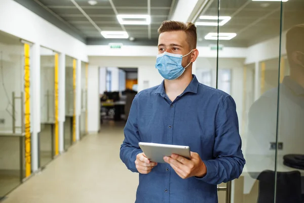 Young man with tablet in his hands standing at the office corridor wearing protective medical mask. Leading business and going to work during coronavirus pandemic. Stay safe concept