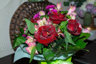 Kırmızı güller ve pembe çiçeklerden oluşan çarpıcı bir düzenleme karanlık bir ahşap masada duruyor. Buket, yemyeşil yapraklara sahiptir ve arka planda dekoratif elementlerle sıcak, rahat bir ev ortamı ile çevrilidir..