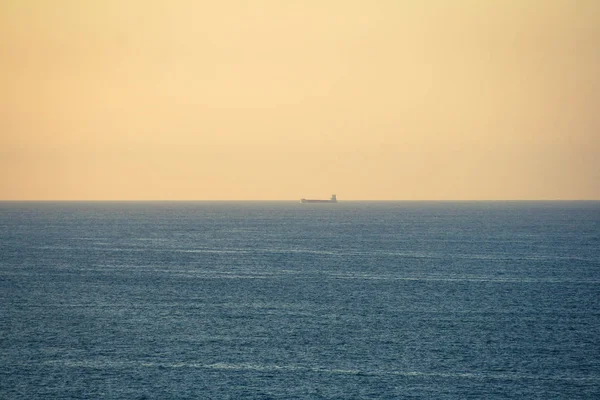 Landscape of an ocean with the horizon line centered on the image, a tanker sailing and an orange sky at sunset