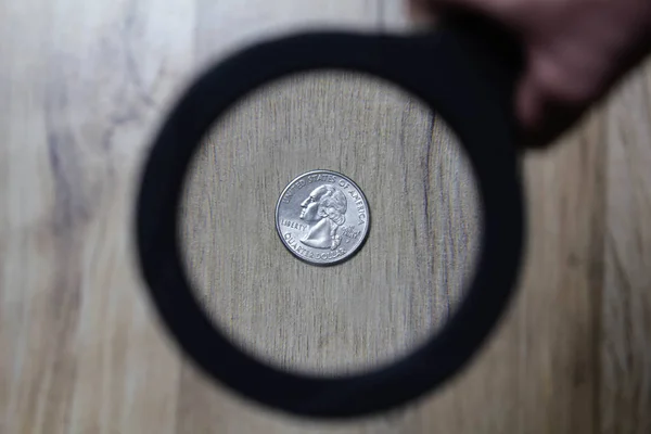 A magnifying glass is aimed at a quarter of a dollar on a wooden parquet