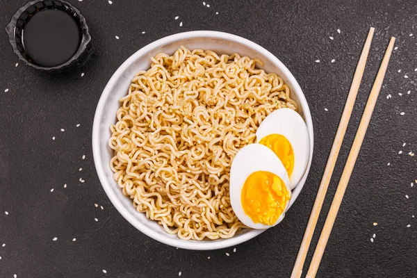 Macarrão em uma chapa com sementes de sésamo e molho de soja, paus de bambu, ovos fervidos em uma mesa preta fecham. Ley plano. espaço de cópia. orientação horizontal — Fotografia de Stock