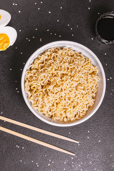 Noedels in een bord met sesamzaad en sojasaus, bamboestokjes, gekookte eieren op een zwarte tafel van dichtbij. Een platte ley. kopieerruimte — Stockfoto