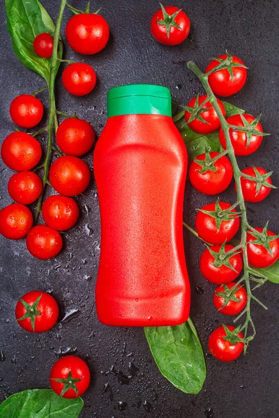 Una bottiglia, un tubetto di ketchup rosso con tappo verde e due rami maturi di pomodorini con spinaci e gocce d'acqua di rugiada su un tavolo nero goffrato. vista dall'alto, posizione piatta, orientamento verticale — Foto Stock